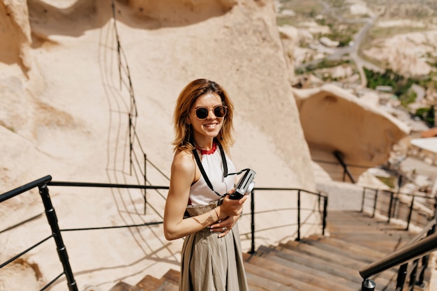 Charming smiling lady with short hairstyle wearing sunglasses holding retro camera and walking among mountains in sunlight