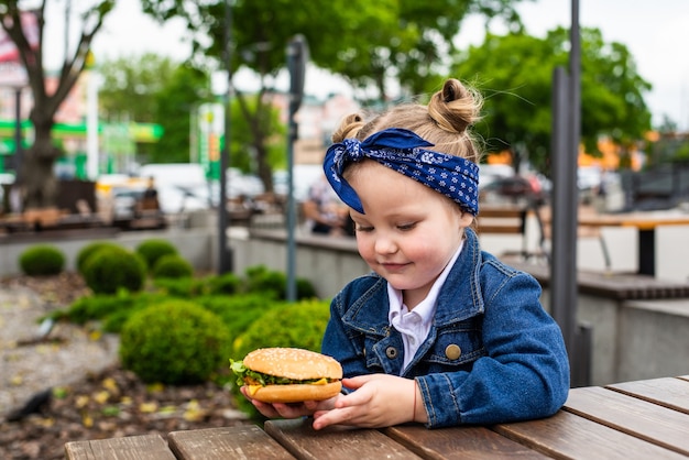 A charming smiling little girle is holding a hamburger in the open air on a sunny day.