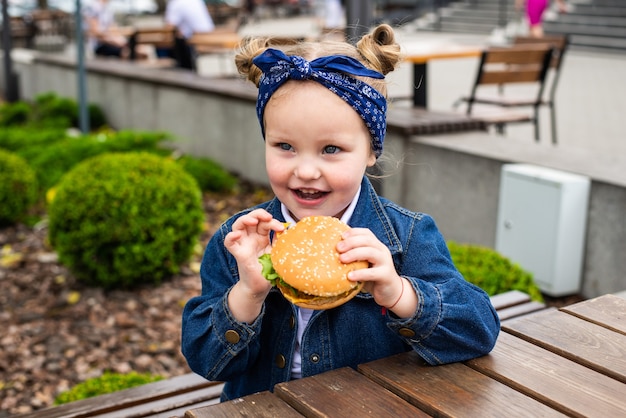 Free photo a charming smiling little girle is holding a hamburger in the open air on a sunny day.