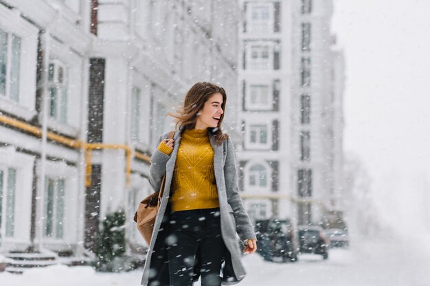Charming smiling young woman in coat with backpack walking in snowfall in Europe city centre. Expressing positivity, true emotions, enjoy snowing, waiting for christmas holidays, smiling to side.