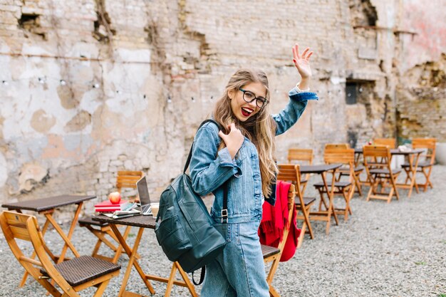 Charming student passed exams perfectly. Adorable girl in a fashionable denim suit leaves the outdoor cafe and says goodbye to friends.