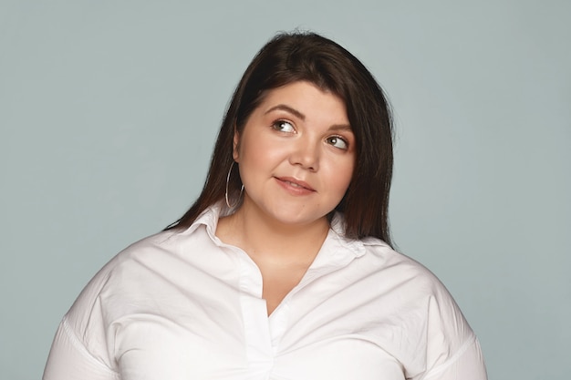 Free photo charming stylish plus size young dark haired female looking up with thoughtful facial expression, daydreaming, thinking of something pleasant. fashionable chubby woman in white shirt posing