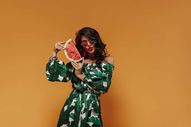 Charming stylish woman with curly long hair with red sunglasses and green and white sundress posing with watermelon and orange backdrop