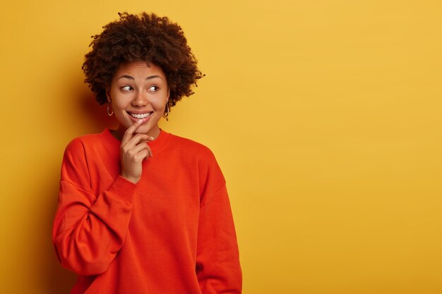 Charming tender curly woman smiles gladfully, looks aside, wears casual clothes, stands against yellow studio wall