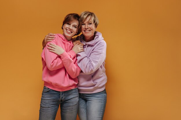 Charming two ladies with lovely smiles and short cool hairstyle in modern pink sweatshirts and trendy jeans hugging on isolated backdrop. 