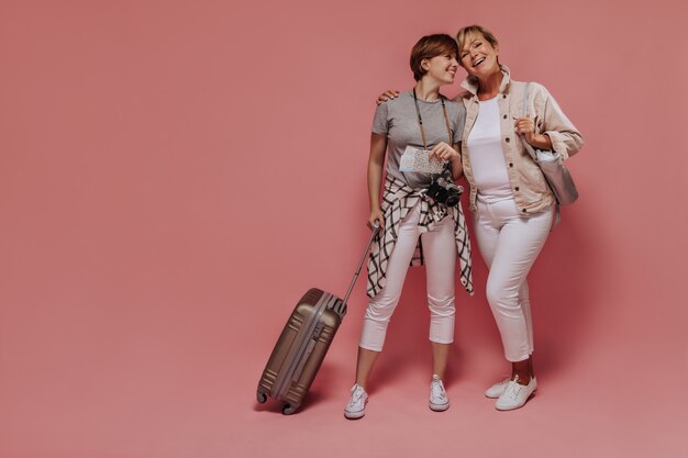 Очаровательные две дамы с короткой крутой прической в легкой современной одежде позируют с билетами, камерой и чемоданом и улыбаются на розовом фоне.