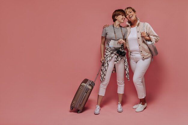 Charming two ladies with short cool hairstyle in light modern clothes posing with tickets, camera and suitcase and smiling on pink background.