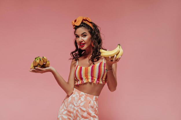 Charming wavy haired girl with orange bandana and earrings in modern bright top and pink trousers looking into camera and holding bananas