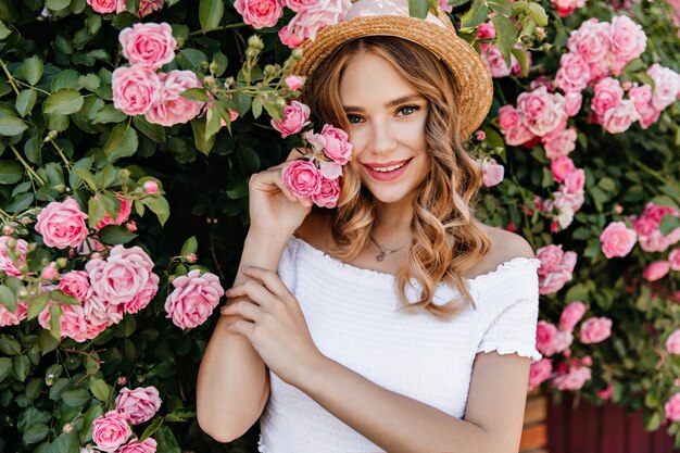 ピンクの花の前に立っている魅力的な白い女性モデル。庭で時間を過ごす流行の帽子のうれしそうな女の子の屋外の肖像画。