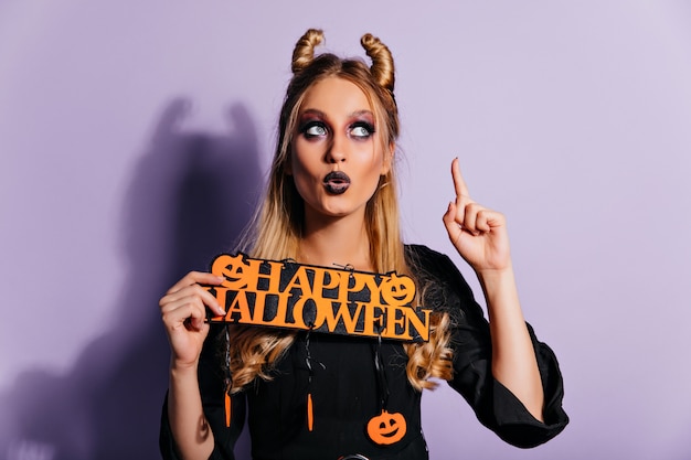 Free photo charming white girl with spooky makeup posing with halloween decor. gorgeous european lady in vampire outfit standing on purple wall.