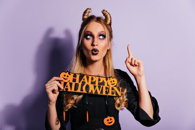 Charming white girl with spooky makeup posing with halloween decor. Gorgeous european lady in vampire outfit standing on purple wall.
