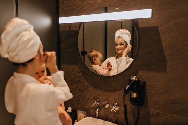 Charming woman in bathrobe puts makeup and holds baby. Mom and daughter observe morning routine in bathroom.