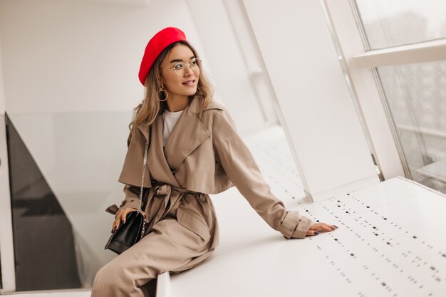 Charming woman in beige coat sits on white windowsill and looks out window