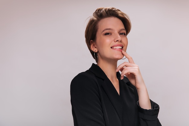 Charming woman in black outfit looks into camera. Attractive short-haired lady in dark jacket smiles and poses on isolated background