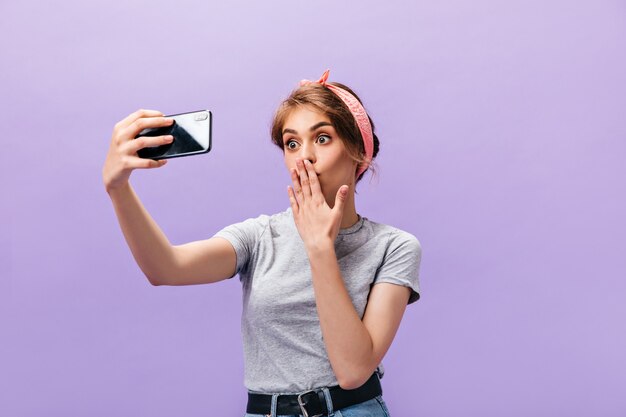 Charming woman blows kiss and takes selfie. Funny beautiful young girl in pink bandana and t-shirt makes photo on isolated background.