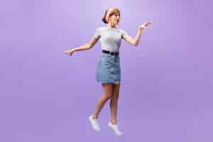 Free photo charming woman in denim skirt jumps on purple background. fashionable pretty girl in pink headband and grey shirt posing.