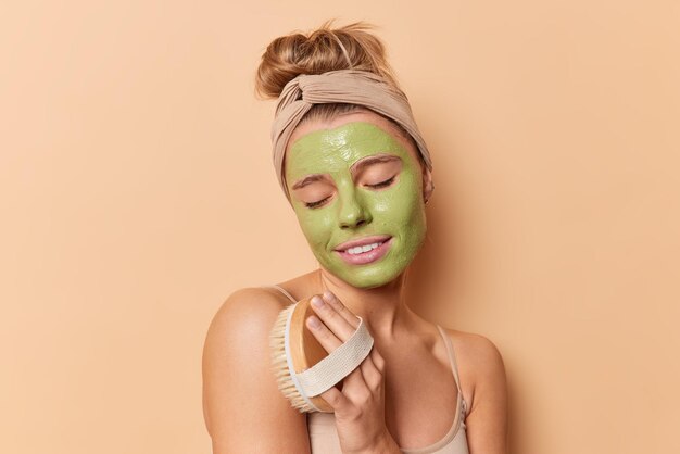 매력적인 여성은 피부 각질을 제거하고 자신이 마른 브러시로 마사지를 하고 녹색 보습 마스크를 착용하고 베이지색 스튜디오 벽에 격리된 맨 어깨에 머리띠를 하고 있습니다. 바디 스크럽