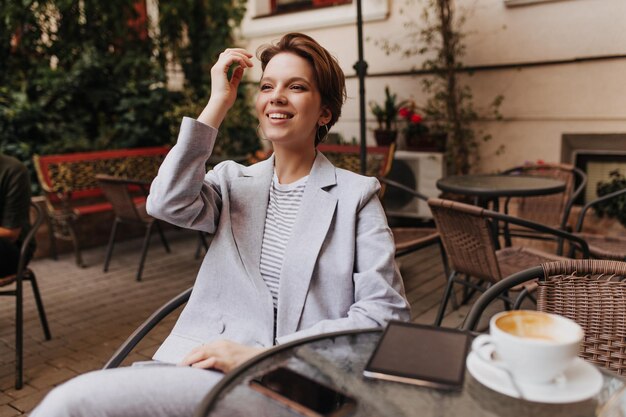 Charming woman in grey suit sitting in cafe Happy woman in oversize jacket smiling and resting in street restaurant