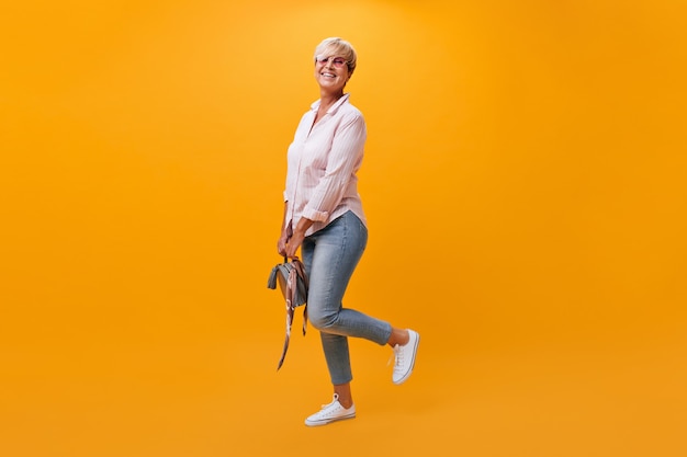 Charming woman in jeans, shirt and sunglasses poses on orange background