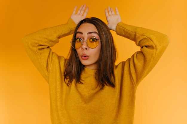 Charming woman making funny faces on bright yellow wall. Carefree caucasian girl in sunglasses fooling around