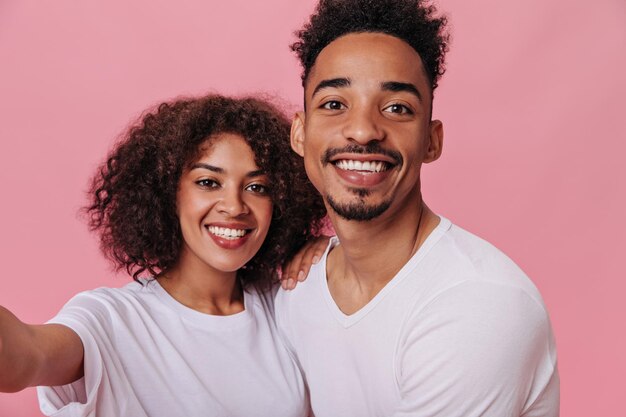 Charming woman and man dressed in white Tshirts smiling and taking selfie Portrait of cheerful curly girl and darkskinned girl in tee posing on pink background