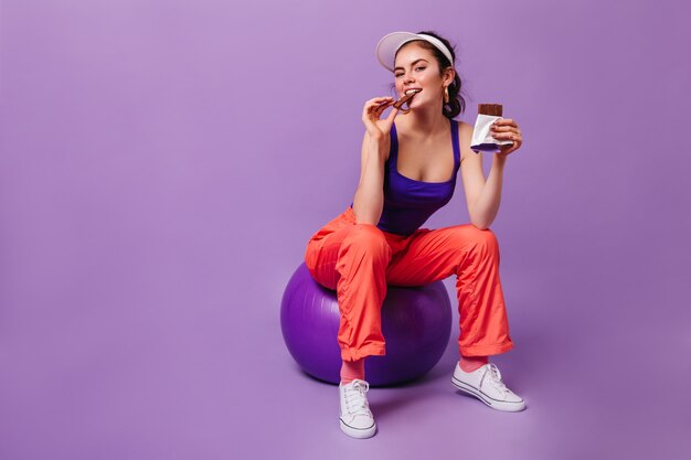 오렌지 스웨트 팬츠와 보라색 탑의 매력적인 여자가 fitball에 앉아 초콜릿을 먹고 있습니다.