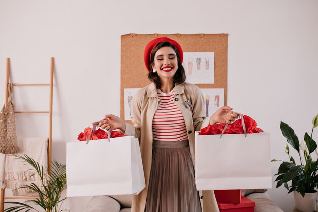 白い買い物袋を保持している赤いベレー帽の魅力的な女性。秋のベージュのコートのポーズで素敵な笑顔で幸せな素敵な女の子。
