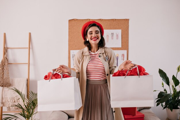 Charming woman in red beret holding white shopping bags. Happy wonderful girl with lovely smile in autumn beige coat posing.