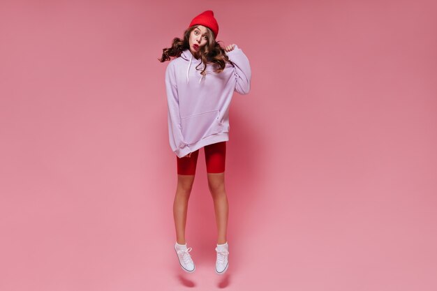 Charming woman in red shorts and oversized purple hoodie jumps on isolated