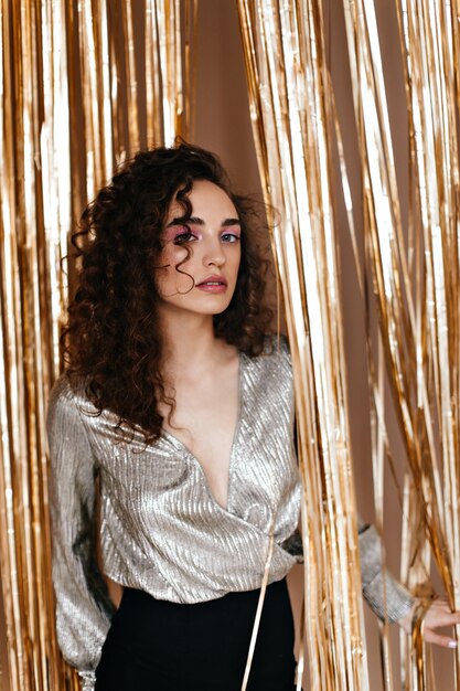 Charming woman in silver outfit looks into camera on festive background