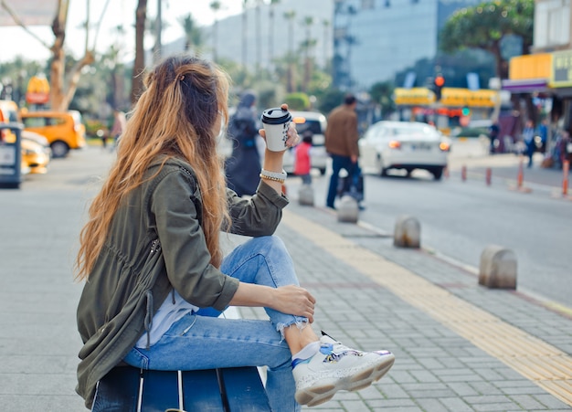 昼間に通りで考えて、コーヒーを保持しているベンチに座っている魅力的な女性。