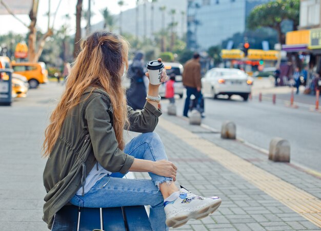 Charming woman sitting on a bench holding coffee, thinking in street during daytime .