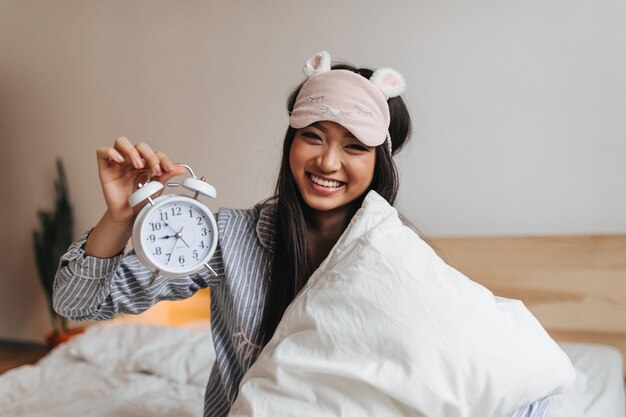 Очаровательная женщина в полосатой пижаме смеется и держит будильник