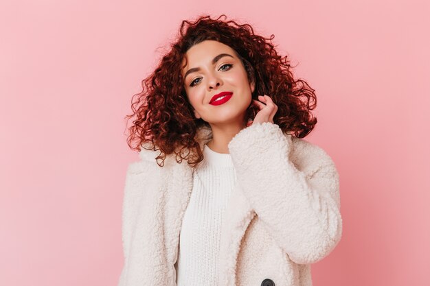 Charming woman in stylish white sweater and fur coat cute smiles. Portrait of curly lady with blue eyes and red lips.