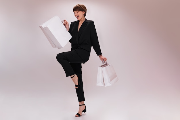 Charming woman in suit moves on isolated background and holds shopping bags. Pretty lady in black jacket and pants jumps with white packages on white backdrop