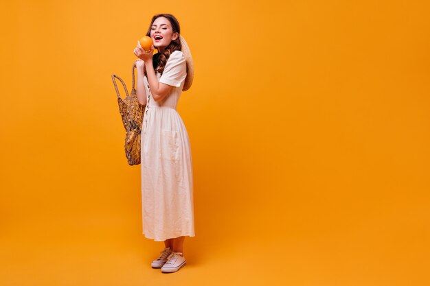 Очаровательная женщина в летнем белом платье держит авоську и кусает апельсин.