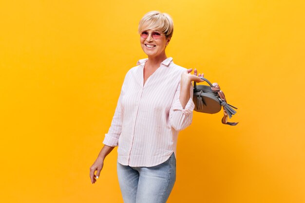 Charming woman in sunglasses and pink shirt posing with handbag on orange background
