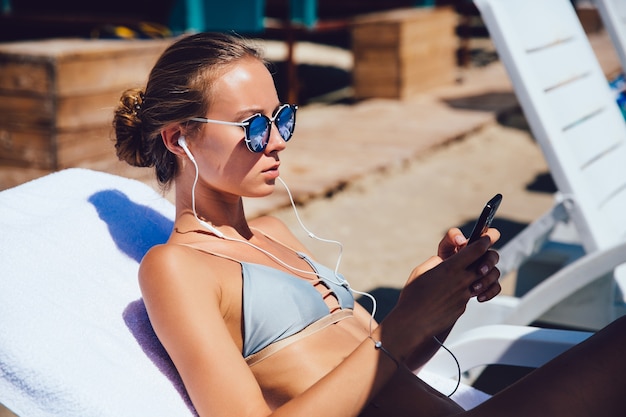 Charming woman in swimsuit, using a mobile phone, in headphones, listening to music