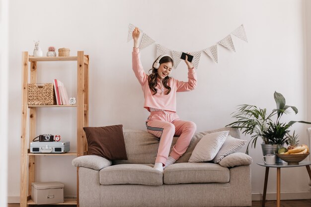 Charming woman in tracksuit dancing, sitting on couch