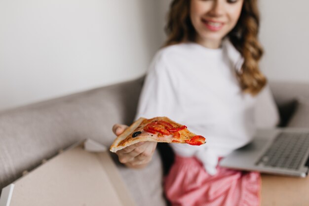 Charming woman using laptop and eating pizza with cheese. Indoor shot of relaxed girl in white t-shirt working with computer and enjoying fast food.