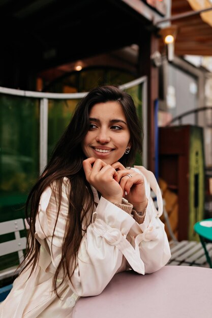 흰 코트를 입은 매력적인 여성이 야외 카페에 앉아 미소를 짓고 거리에서 기분 좋게 걷고 있다