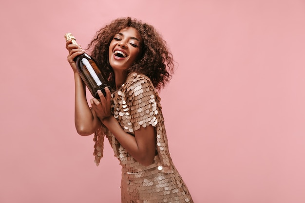Очаровательная женщина с вьющейся прической брюнетки в блестящем стильном платье смеется и держит бутылку с вином на изолированной стене.