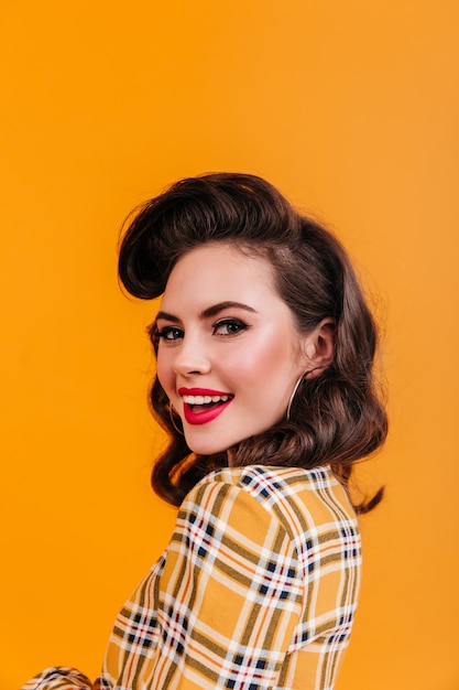 Free photo charming woman with curly hairstyle looking over shoulder. smiling pinup girl in checkered shirt posing on yellow background.