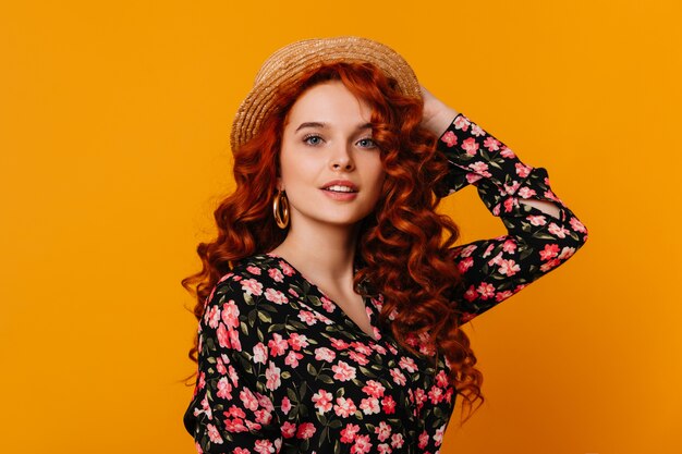 長い赤い髪の魅力的な女性がカンカン帽をかぶっています。オレンジ色のスペースに美しいシルクのブラウスを着た巨大な金のイヤリングを持つ女性。