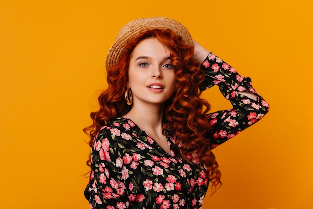 Charming woman with long red hair puts on boater. lady with massive gold earrings dressed in beautiful silk blouse on orange space.