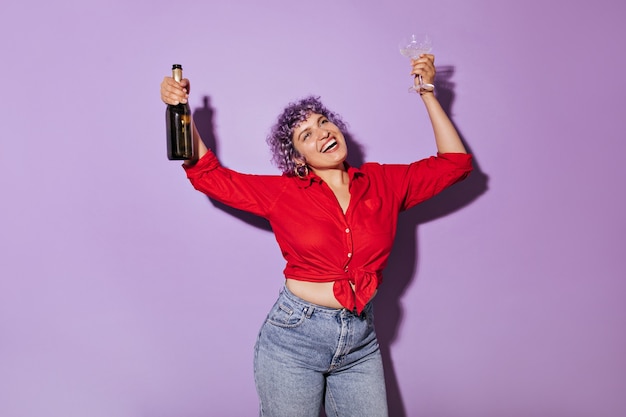 Charming woman with purple hair in shirt and jeans laughs with glass in his hand. Wonderful lady in bright clothes holds bottle of wine.