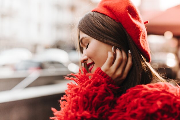 Charming woman with red lipstick is smiling with her eyes closed. woman in red warm outfit touches her silver earrings.