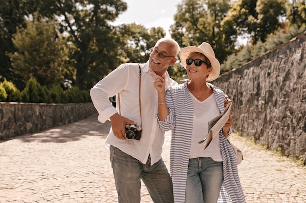 Charming woman with short hairstyle in hat and blue blouse holds map, points to side and smiles with grey haired man with camera in park.