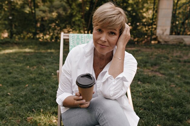 Charming woman with short hairstyle in white shirt and jeans holding cup of coffee, looking into camera and sitting in park.