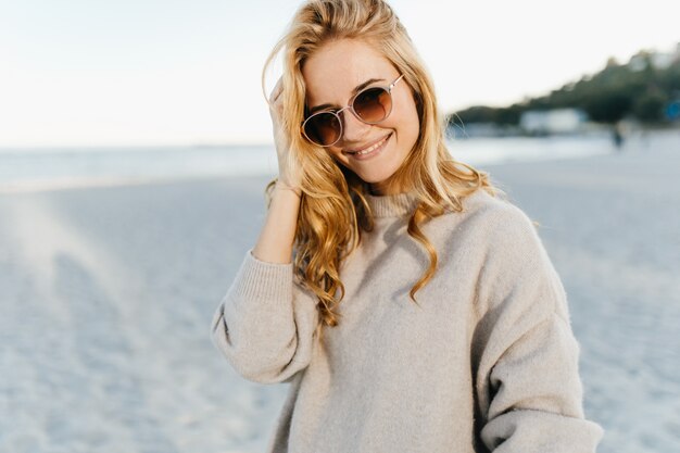 Charming woman with wavy blind hair, dressed in light sweater and sunglasses with smile against sea.